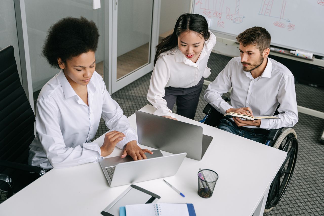 A diverse team of professionals working together in an office with laptops and a wheelchair user.