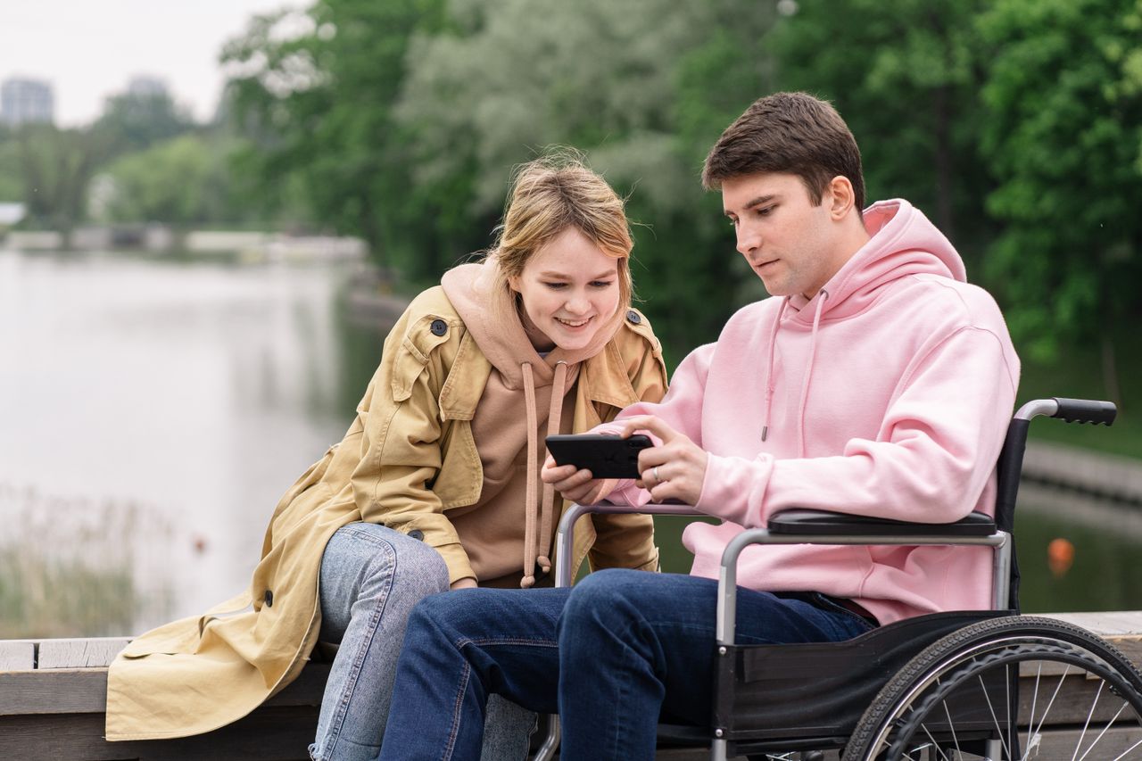 A couple sitting by a lake, looking at a smartphone. One is in a wheelchair, promoting inclusivity.
